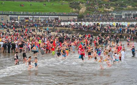 a photo of the event showing hundreds of people in fancy dress running into the water as an audience watches from the beach